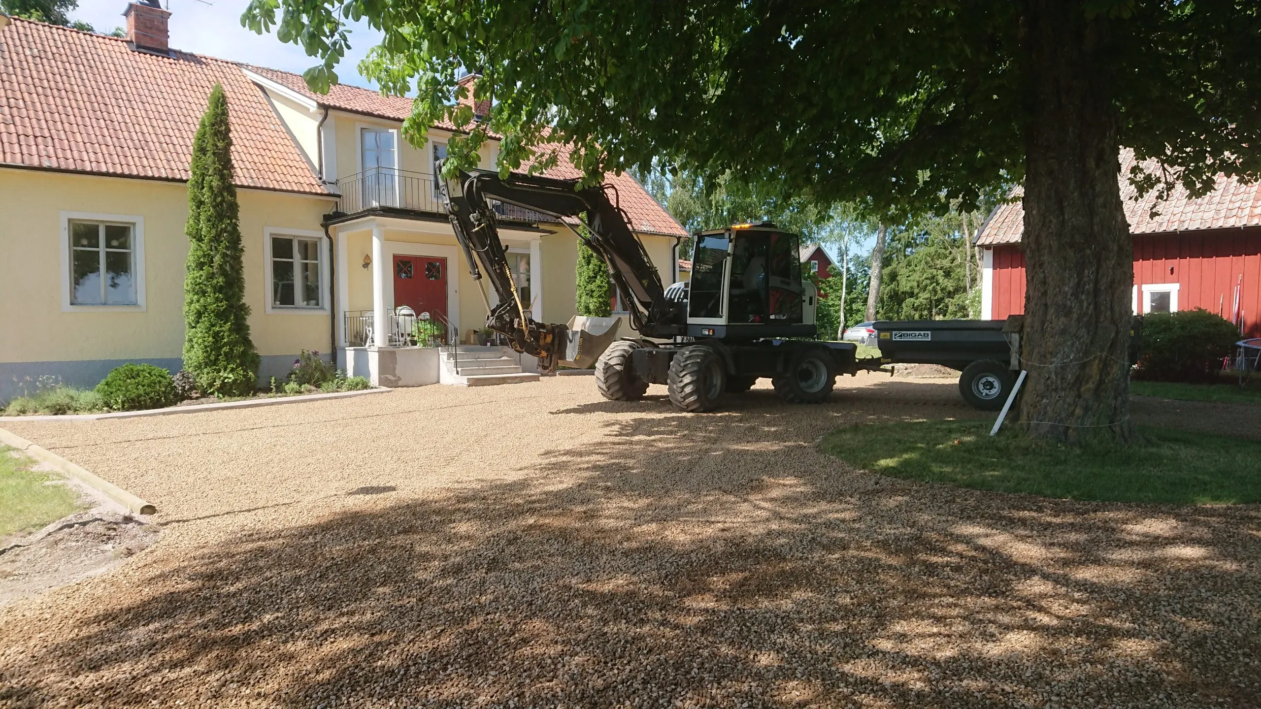 Hjulgrävare med vagn på en grusplan framför ett gult bostadshus
