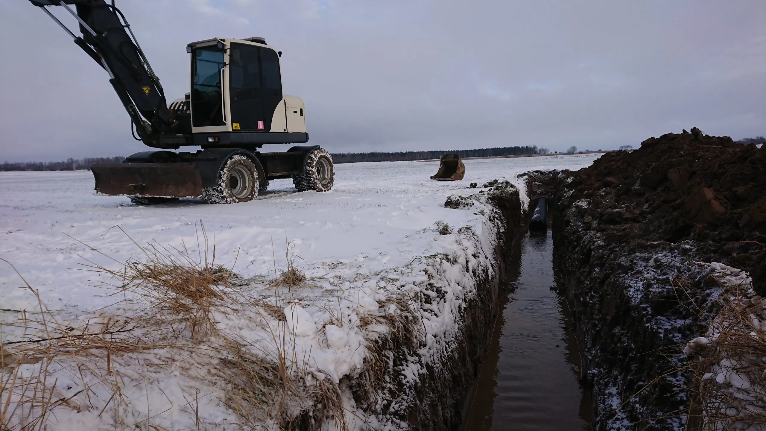 Grävmaskin på ett snötäckt fält bredvid ett nygrävt dike